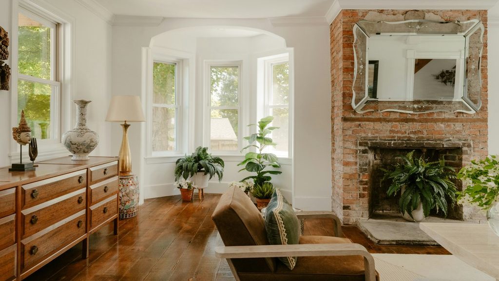 Bright family room with a brown leather chair, wood dresser, bay windows, exposed brick fireplace, and leafy indoor plants.