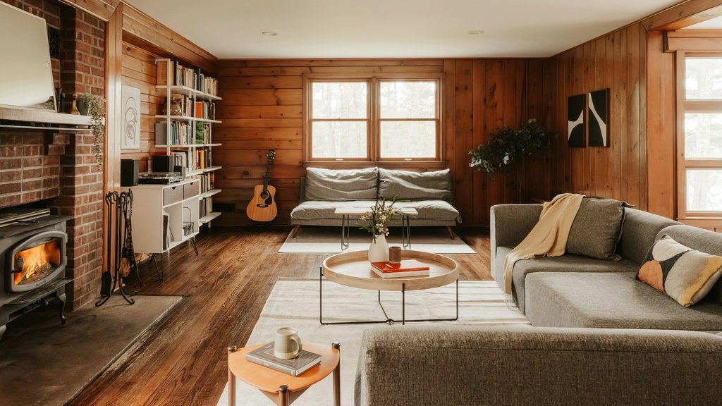 Cozy wood-paneled family room with a fireplace, gray sofas, layered rugs, round coffee table, bookshelf, and warm natural light.