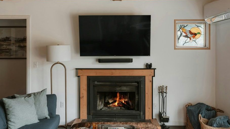 Living room with a fireplace below a wall-mounted TV, wood ceiling, blue sofa, floor lamp, coffee table, and woven storage baskets.