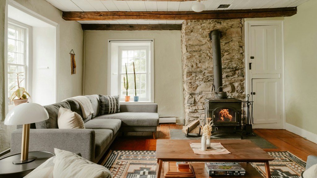 Cozy family room with a gray sectional, stone fireplace, wood-burning stove, exposed ceiling beams, patterned rug, and wood coffee table.