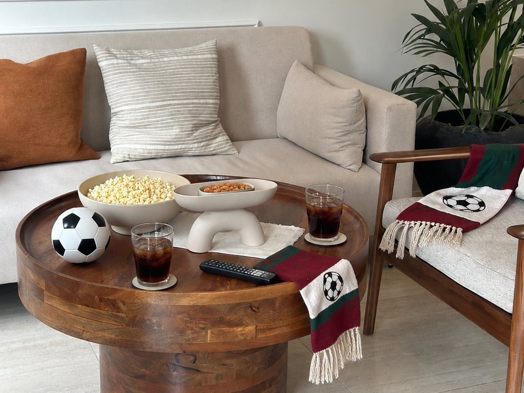 Round wooden coffee table with popcorn, drinks, a remote, soccer ball, and team-colored scarves arranged for a cozy World Cup watch party.