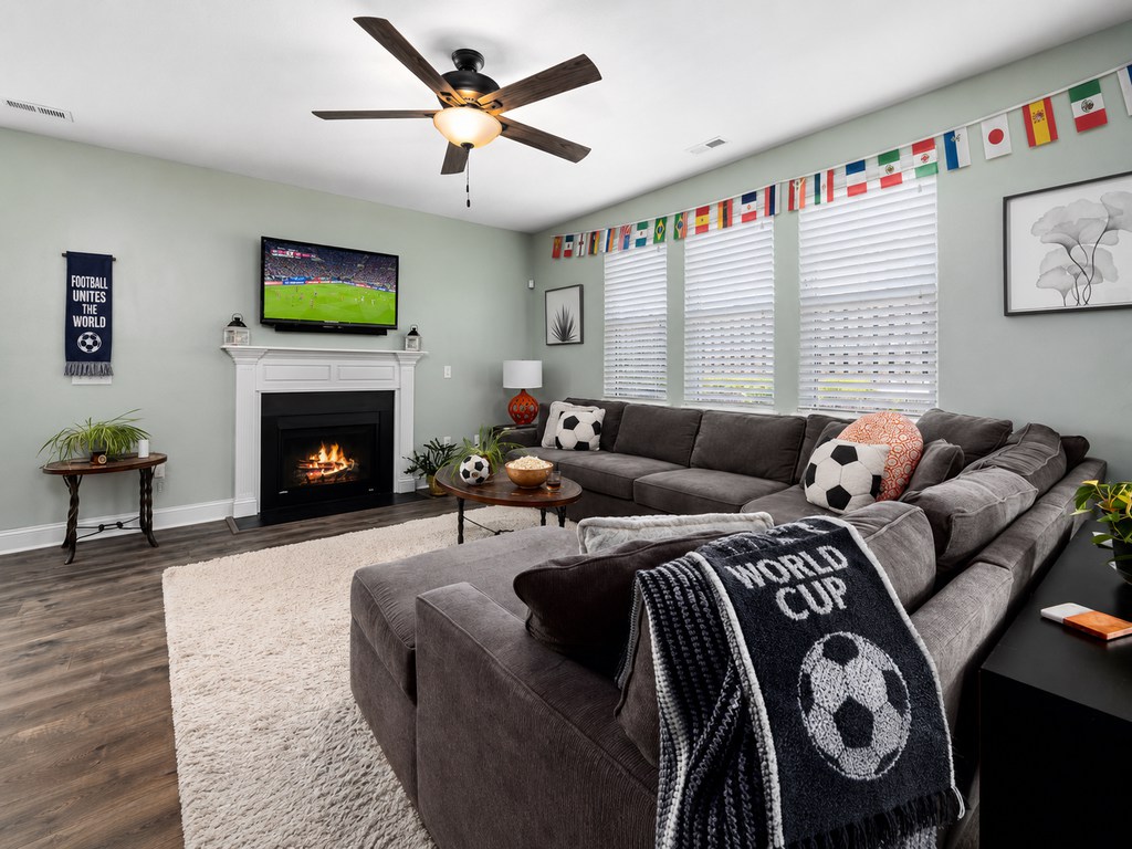 Living room with a gray sectional sofa facing a TV showing a soccer match, decorated with World Cup flags, soccer pillows, blanket, and snacks.
