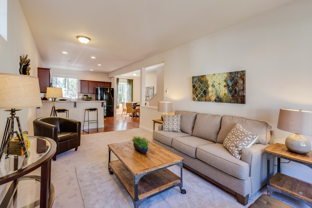 Bright open-plan living room with beige sofa, leather armchair, wood coffee table, side lamps, and wall art creating asymmetrical balance.