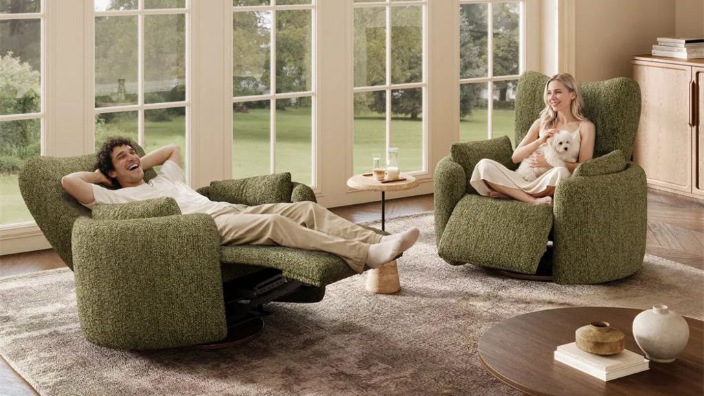 Two green swivel recliner chairs in a sunlit living room, with one person reclining and another seated with a small white dog beside a round wood coffee table.