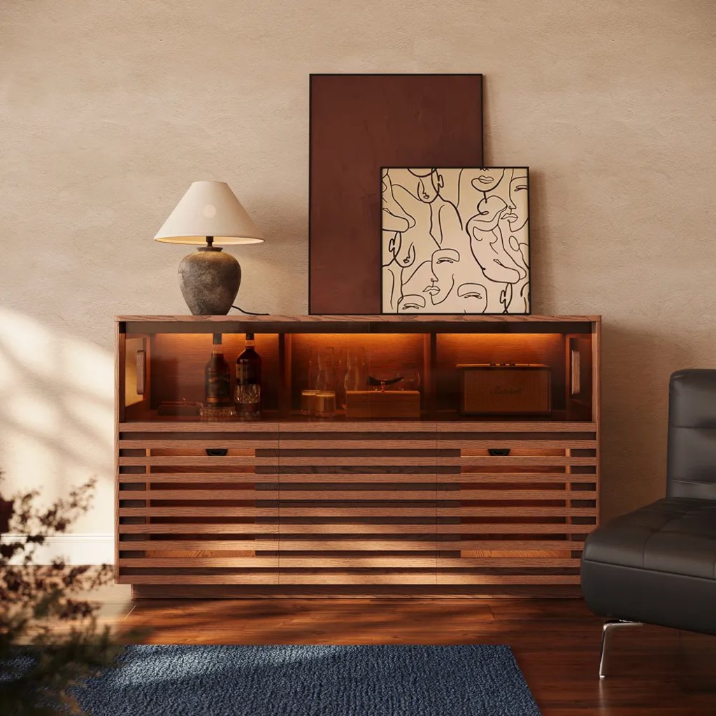 Warm wood buffet cabinet with glass display shelves, a table lamp, framed artwork, bottles, and a leather chair in a modern living room.
