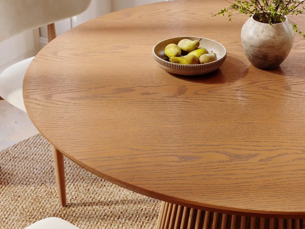 Close-up of a round oak dining table with a bowl of pears and a ceramic vase in a bright dining area.
