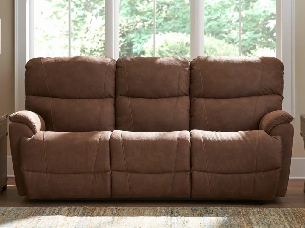 Brown three-seat reclining sofa with plush cushions and padded arms placed in front of large sunlit windows in a cozy living room.