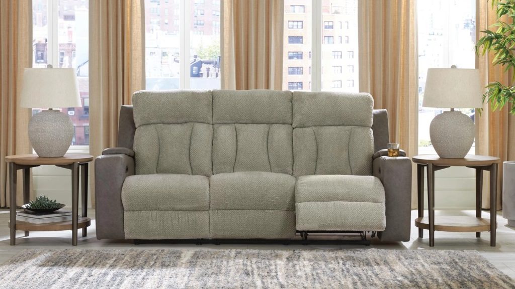 Light gray dual reclining loveseat with one footrest extended, framed by matching side tables and lamps in a bright apartment living room.