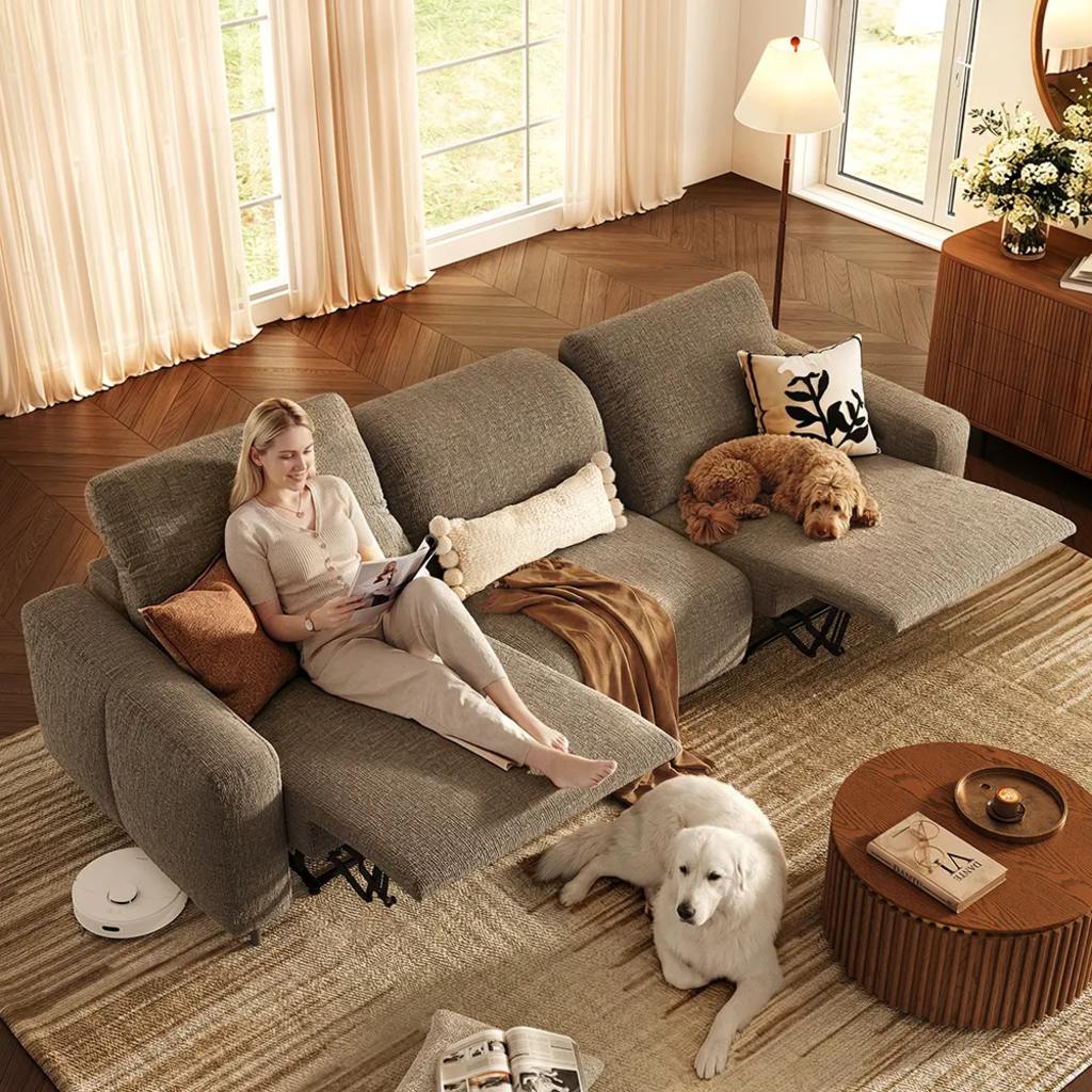 Overhead view of a woman relaxing on a gray reclining sectional with two dogs, a round wood coffee table, and a robot vacuum in a sunlit living room.
