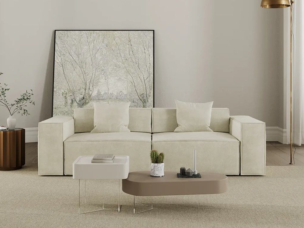 Minimalist living room with a cream modular sofa, layered coffee tables, a neutral rug, and soft brass floor lighting.