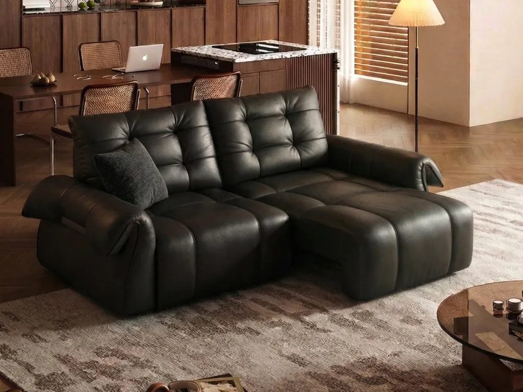Black leather power sofa bed with two seats extended in a warm open-plan living room and kitchen.