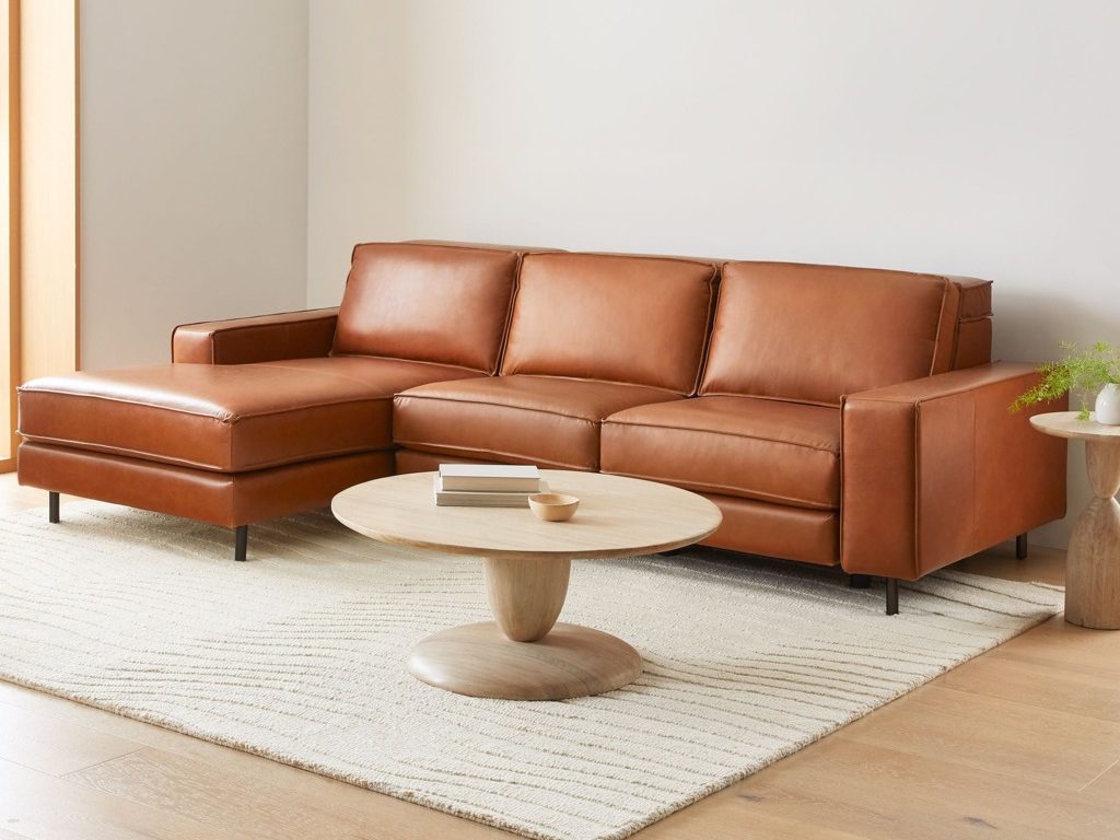 Cognac leather chaise sectional sofa with slim black legs in a warm modern living room, styled with a round wood coffee table and textured neutral rug.