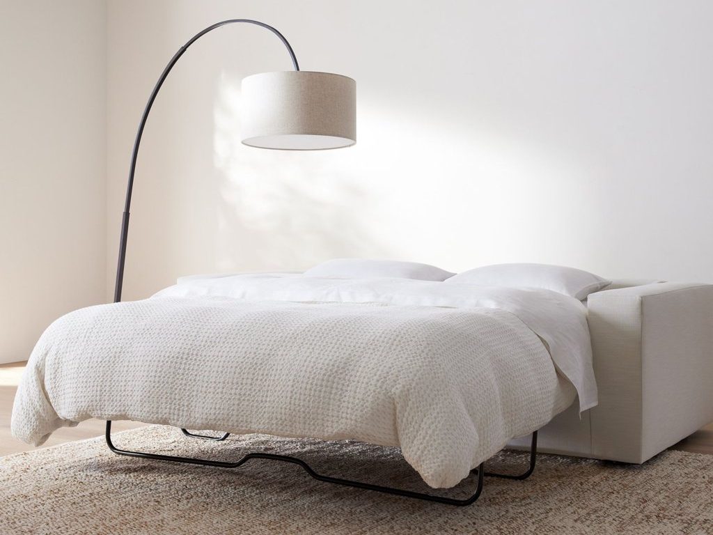 White sleeper sofa unfolded into a bed with pillows and a textured blanket in a minimalist room.