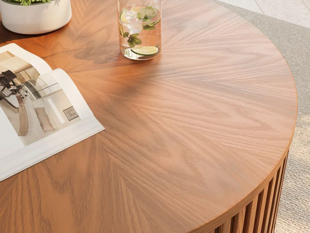 Close-up of a round light wood coffee table with a glass carafe, potted plant, and open design magazine in a sunlit living room.