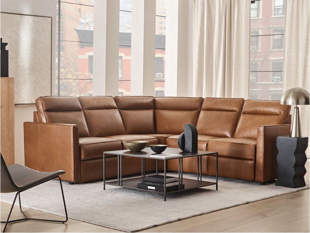 Brown leather reclining sectional sofa in a bright modern apartment living room with tall windows, sheer curtains, a black metal coffee table, and neutral decor.