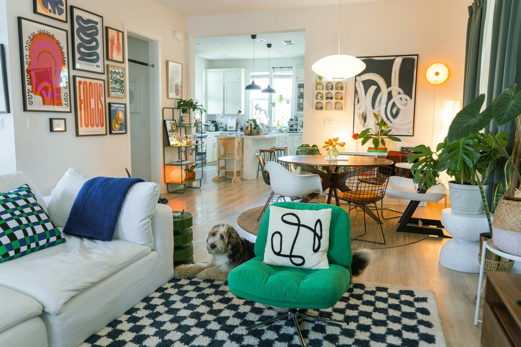 Eclectic open-plan living and dining room with colorful wall art, green accent chair, patterned rug, white sofa, plants, and layered lighting.