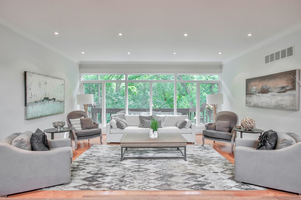 Bright living room with gray sofas, white couch, classic accent chairs, large patterned rug, wood coffee table, and wide windows overlooking greenery.