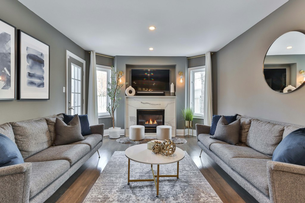 Living room with two gray sofas facing a marble fireplace and wall-mounted TV, centered around a white coffee table and patterned area rug.