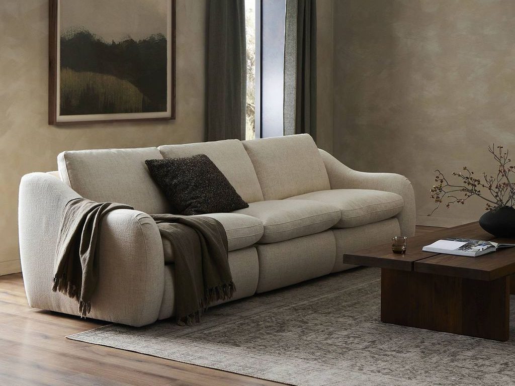 Cream motion reclining sofa with rounded arms and deep cushions in a warm, modern living room with dark curtains, abstract wall art, and a wood coffee table.