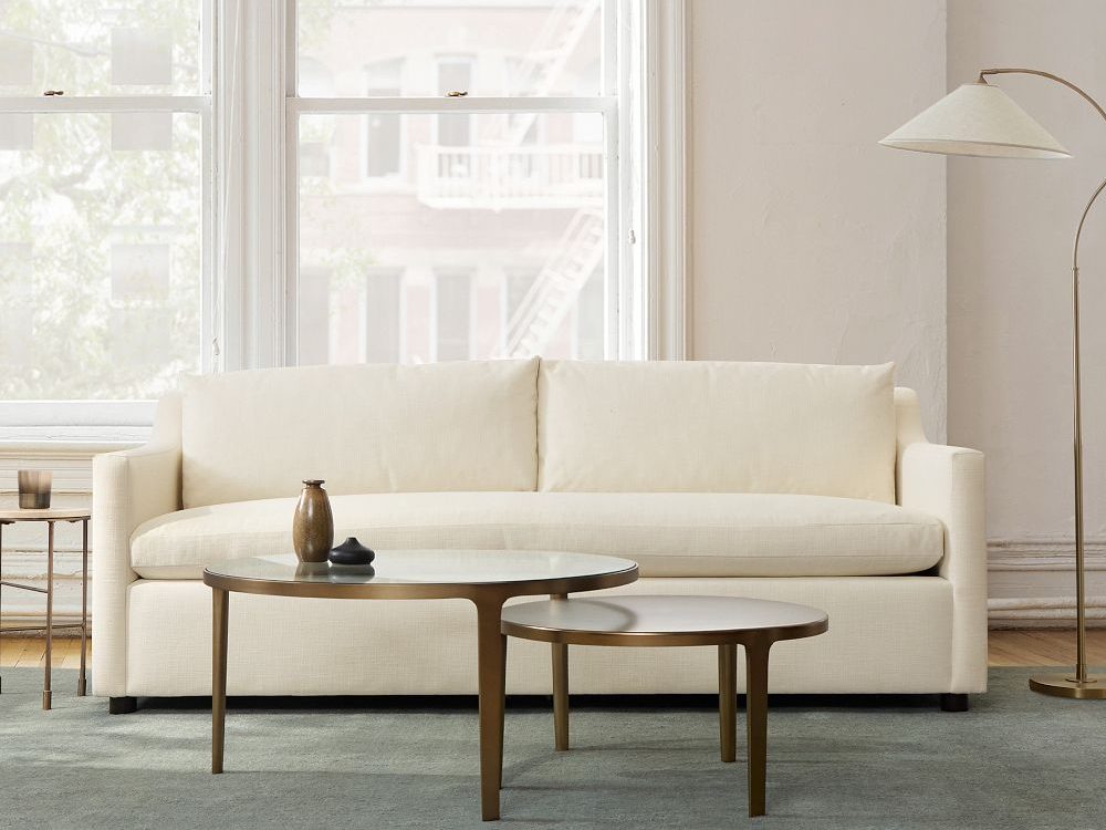 West Elm Marin Slope Arm Queen Sleeper Sofa in beige color near a glass top coffee table, a side table with a cup on top and a floor lamp in a bright apartment
