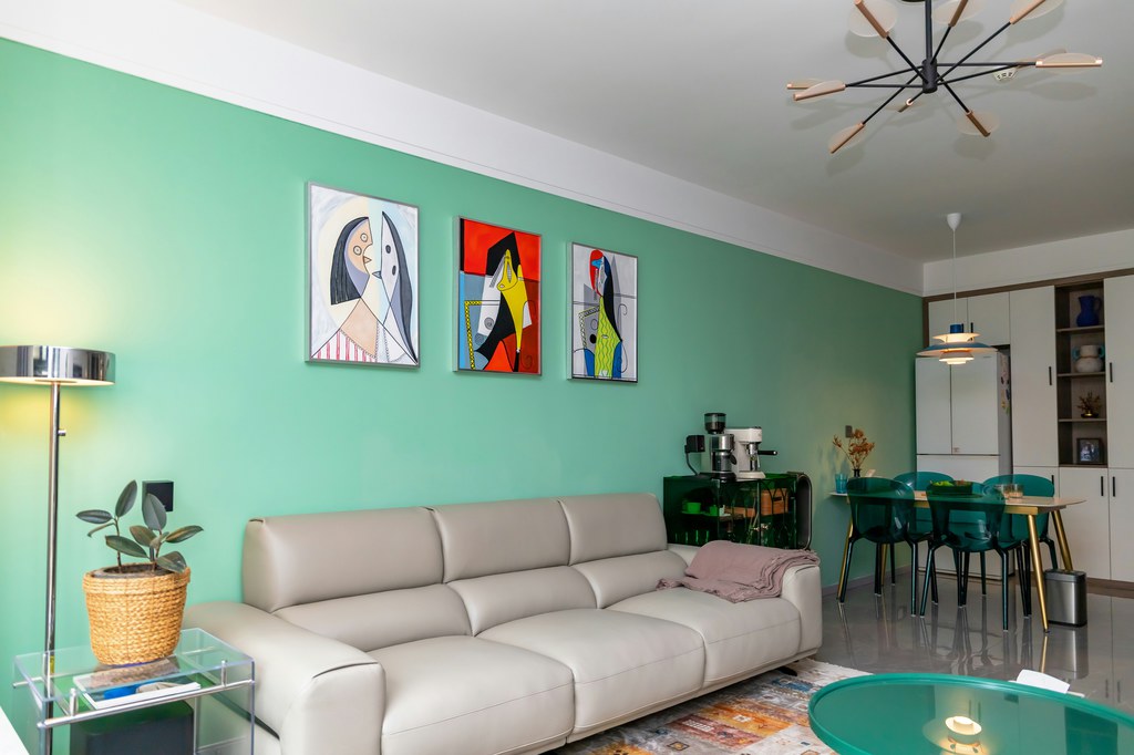 Eclectic living room with mint green accent wall, cream sofa, colorful abstract art, glass tables, modern lighting, and open dining area.