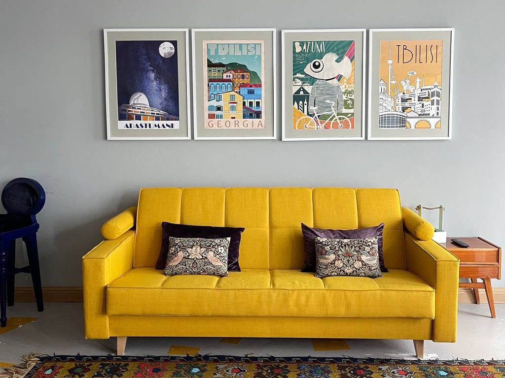 Mustard yellow sleeper sofa with patterned pillows in a colorful living room, framed travel art above and a red geometric rug below.