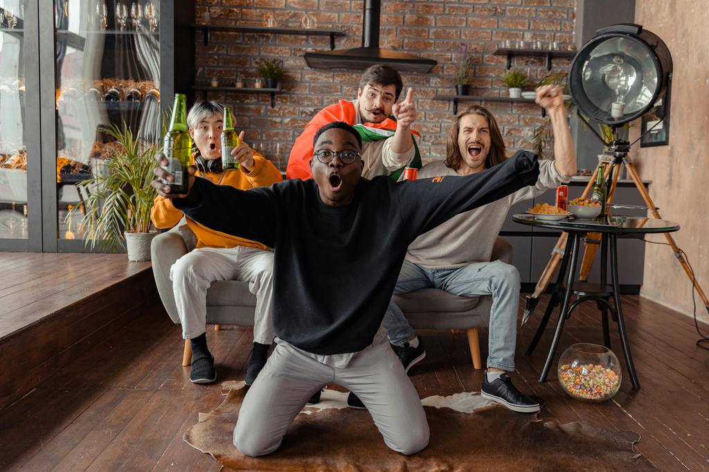 Group of friends cheering in a small living room while watching a soccer match, with snacks, drinks, and casual seating for a watch party.