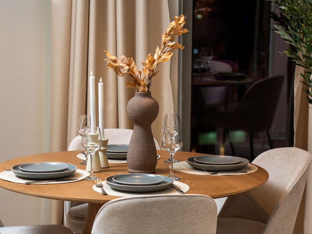Round wood pedestal dining table with upholstered chairs, neutral place settings, tall white candles, and a ceramic vase with dried branches.