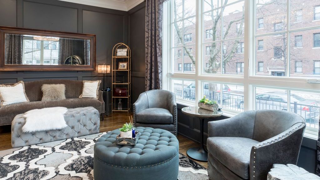 Gray upholstered accent chairs and ottomans arranged in a dark, elegant living room with a sofa, patterned rug, and large windows.