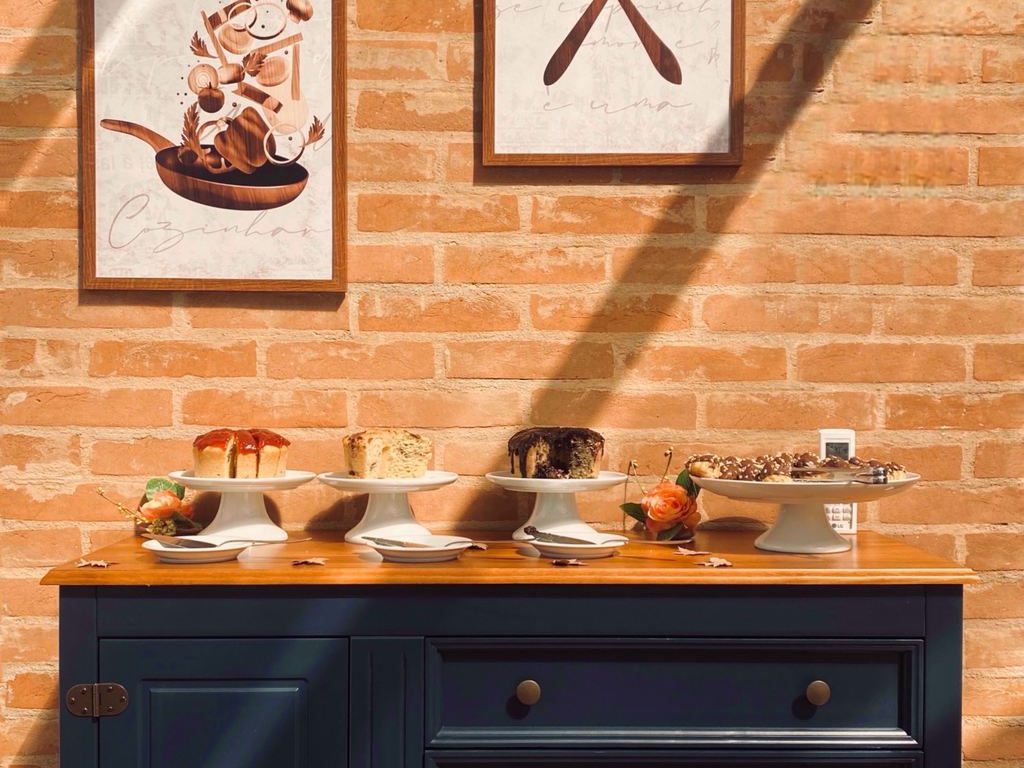 Dark blue buffet cabinet topped with cakes and dessert stands beneath framed kitchen-themed artwork on an exposed brick wall.