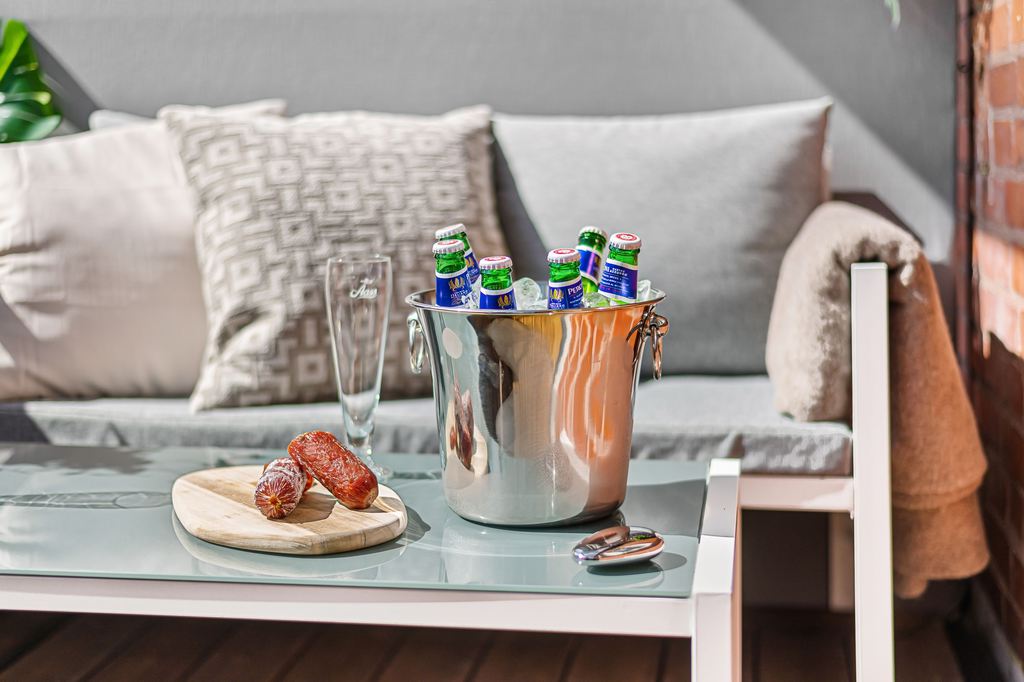 Glass coffee table holding bottled drinks in an ice bucket, a glass, and sliced sausage beside a light gray outdoor sofa.