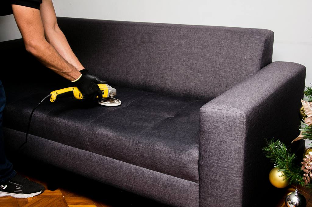 a man with gloves is cleaning a grey fabric sofa with cleaning machine in his living room