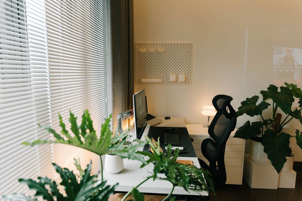 Modern home office with indoor plants around a desk, large window blinds, office chair, and warm lighting for a natural workspace.