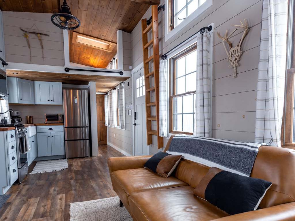 Modern rustic living room with brown leather sofa, wood ceiling, white shiplap walls, large windows, woven rug, and open kitchen layout.