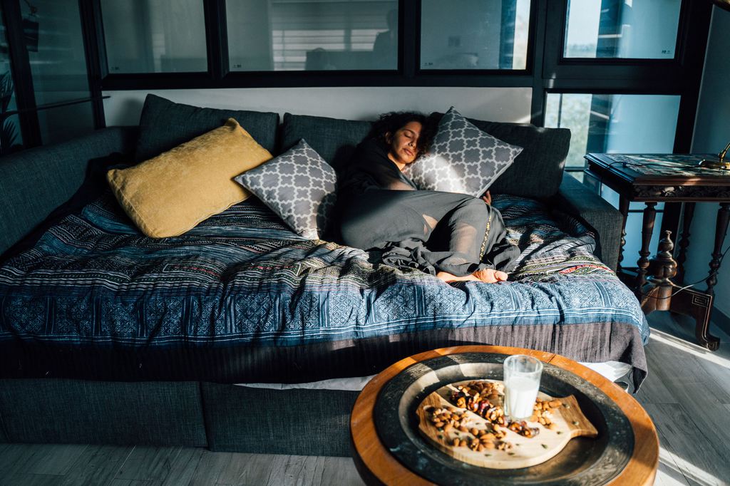 Woman resting on a large sofa bed with layered pillows and a patterned blanket in a modern living room.