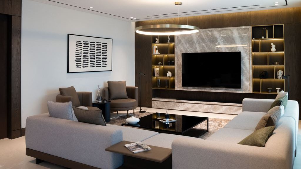 Contemporary living room with two light gray sofas, accent chairs, a black coffee table, circular pendant light, and a built-in TV wall with illuminated shelves.
