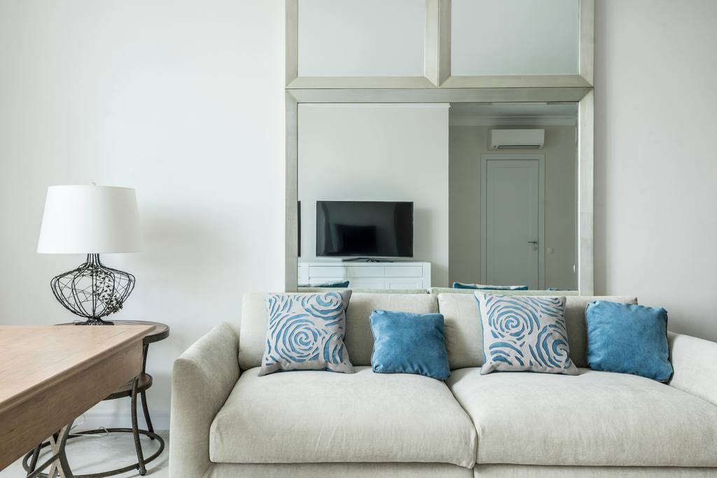 Light beige sofa with blue throw pillows in a bright living room, styled beside a wooden desk, white lamp, and wall mirror.