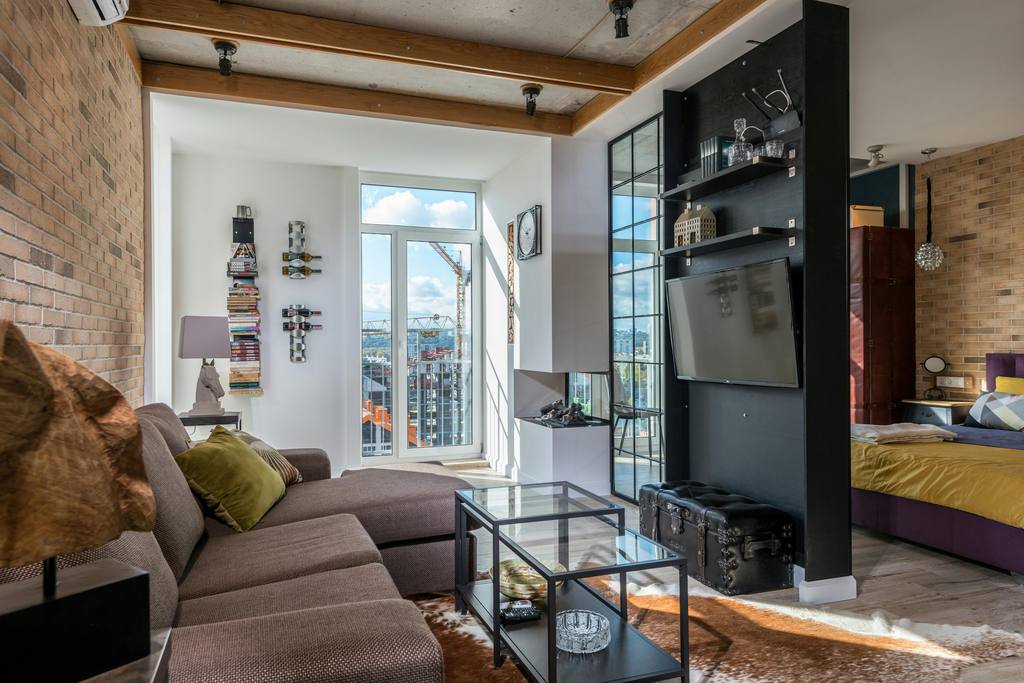 Long narrow open living room with sectional sofa, glass coffee table, wall-mounted TV, and adjacent bedroom visible through a modern partition.