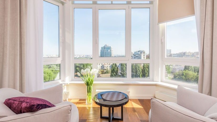 Bright sitting area with two curved armchairs facing a round side table beside large bay windows, neutral curtains, roller shades, and a city view.
