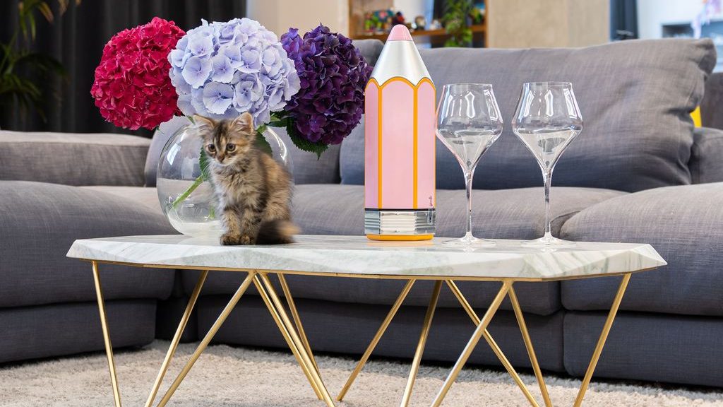 White marble coffee table with gold metal legs, styled with hydrangeas, glassware, a decorative bottle, and a kitten in front of a gray sofa.