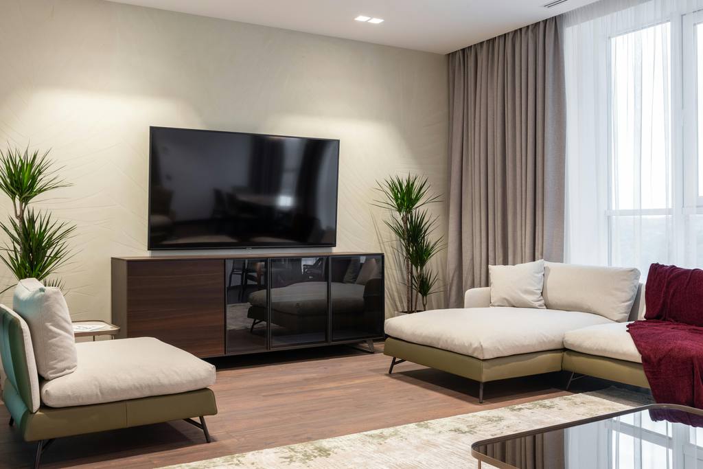 a modern living room with an accent chair, a sofa with chaise, a glass coffee table, a wood media console with glass slide doors and a standing tv between two plants