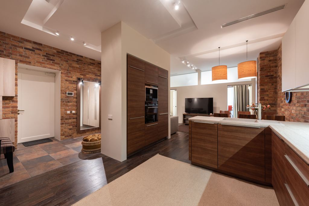 Open-plan warm modern interior with wood kitchen cabinets, brick walls, pendant lights, entryway mirror, and connected living area.