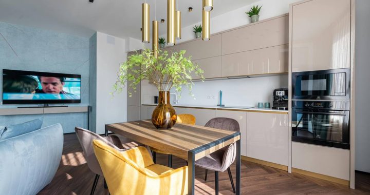 Open-plan dining area with a rectangular wood dining table, colorful upholstered chairs, pendant lights, built-in kitchen cabinets, and nearby living room space.