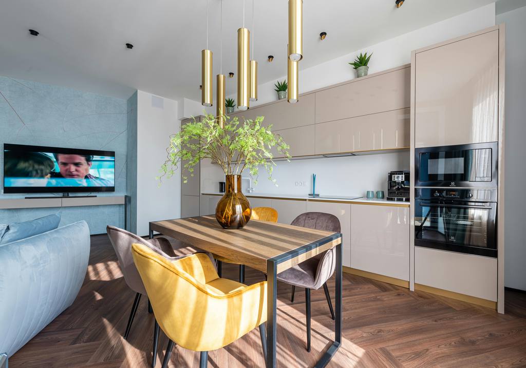 Open-plan dining area with a rectangular wood dining table, colorful upholstered chairs, pendant lights, built-in kitchen cabinets, and nearby living room space.