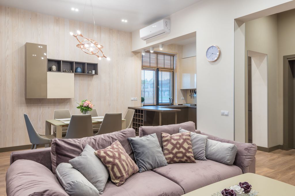Open-plan transitional living and dining room with a mauve sectional sofa, neutral dining set, wood flooring, pendant lighting, and soft patterned pillows.