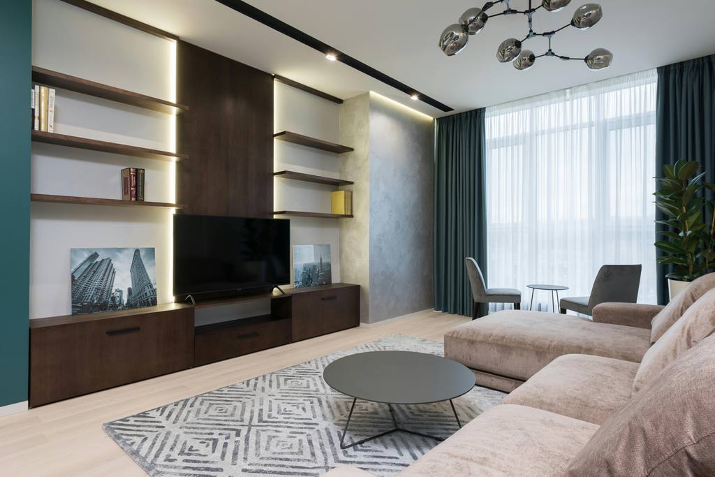 a long and narrow living room with a sofa with chaise, a round coffee table, a tv on a wood media console, with two chairs and a round small desk on end as a reading area