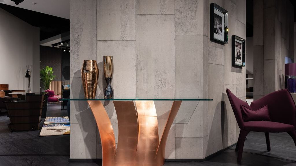 Glass-top console table with a sculptural copper base, styled with metallic vases against a textured gray wall.
