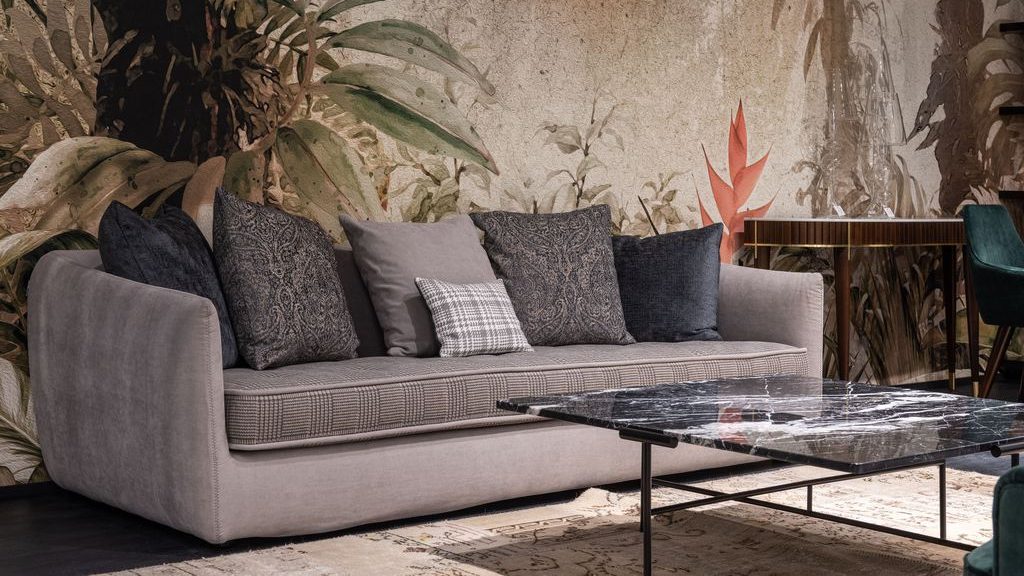 Gray upholstered sofa with layered cushions in a modern living room, set against a botanical mural and marble coffee table.