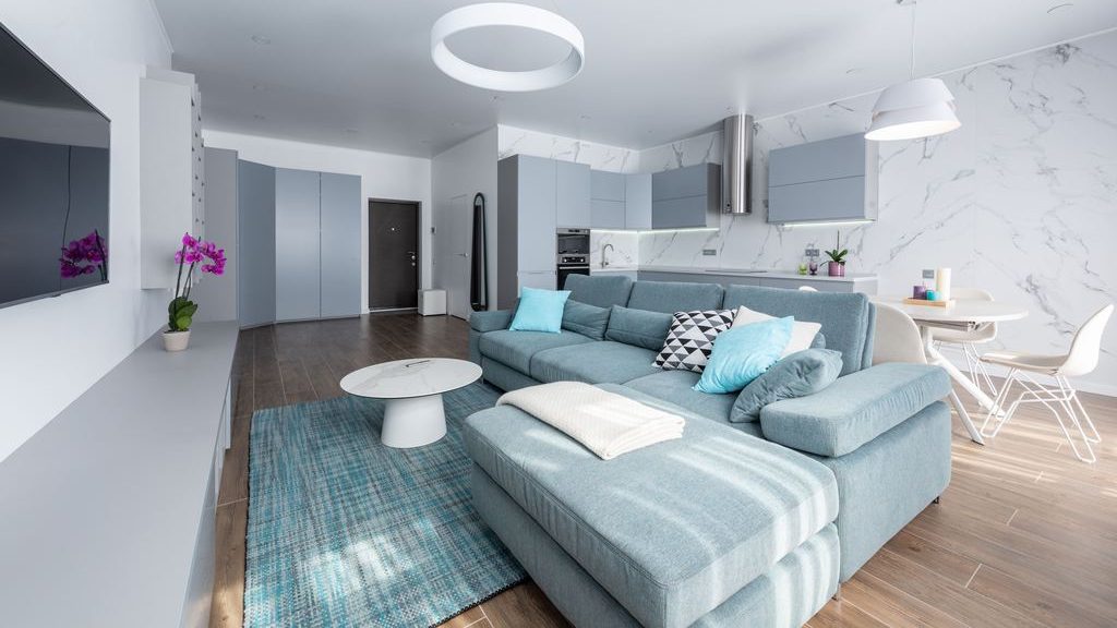 Modern open-concept living room with a blue sectional sofa, chaise lounge, round coffee table, and white kitchen and dining area.