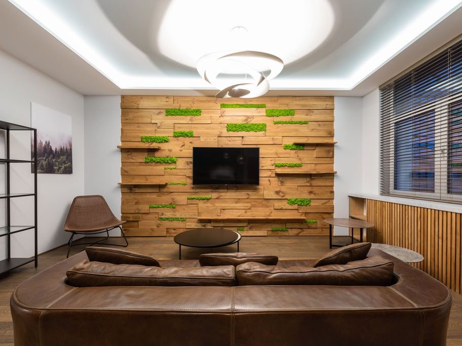 Brown leather sofa facing a wall-mounted TV in a modern living room with wood paneling, greenery accents, and warm lighting.